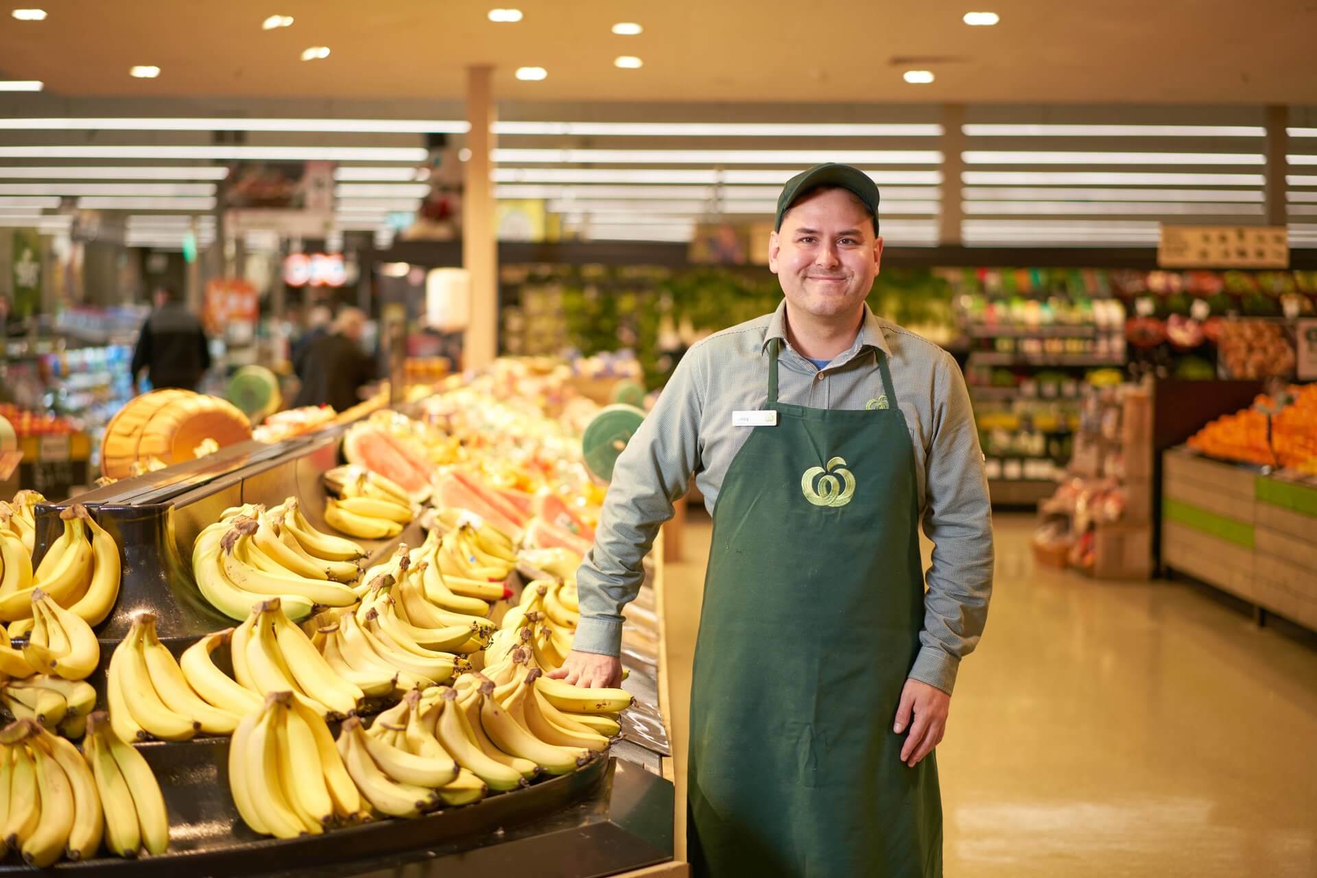 Caley smiles in his Woolworths uniform next to fresh bananas