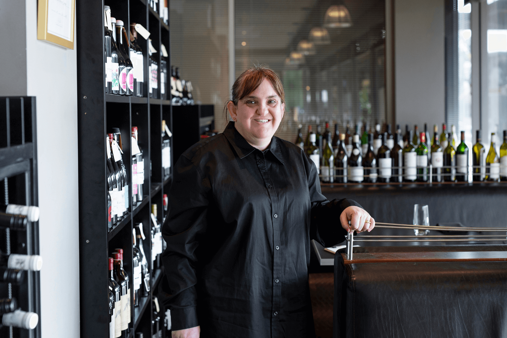 Tammy smiles as she stands next to a wall of wine bottles and a bar behind her