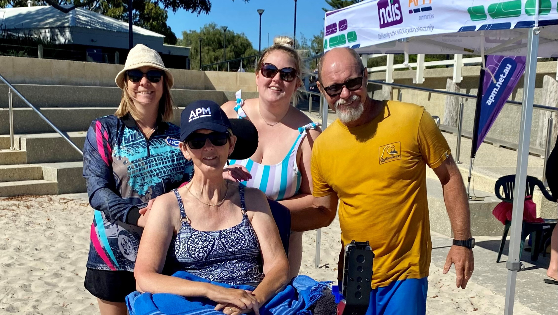 4 people at the beach smiling at the camera, one of them is in a wheelchair