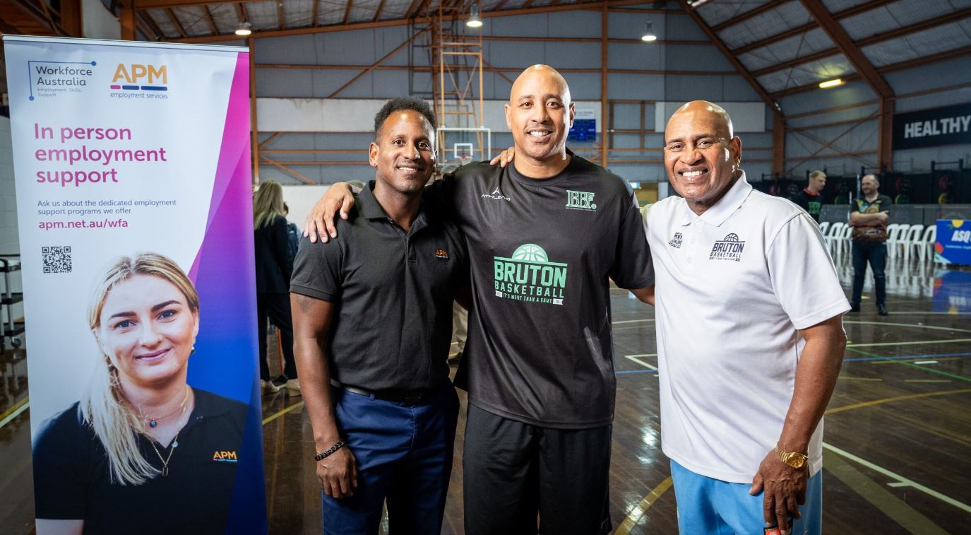 Three men stand with linking arms around shoulders next to an APM banner