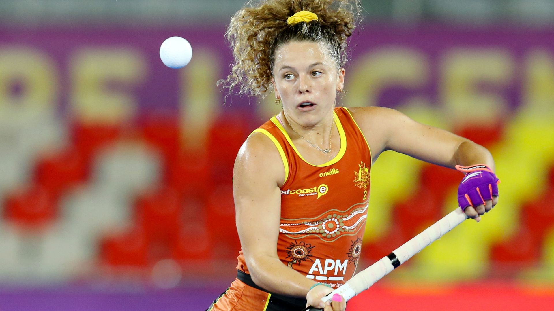 Australian women’s hockey team player wearing an orange Indigenous design uniform in action, holding a hockey stick and focusing on the ball. APM logo is visible on the uniform. Colorful stadium seating in the background.