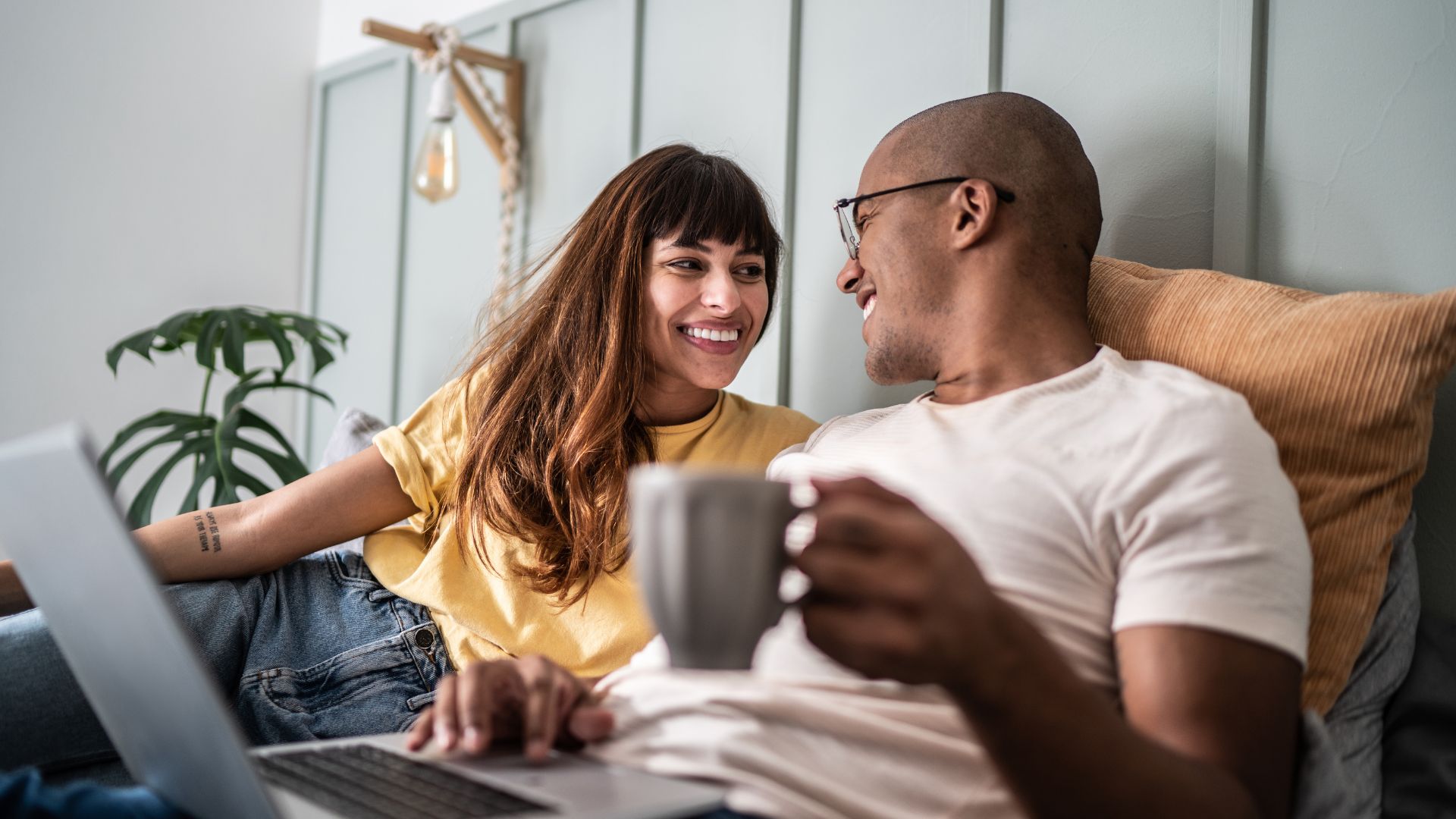  A couple relax on a couch at home, smiling at each other while using a laptop, with one person holding a mug in a cosy living room setting.