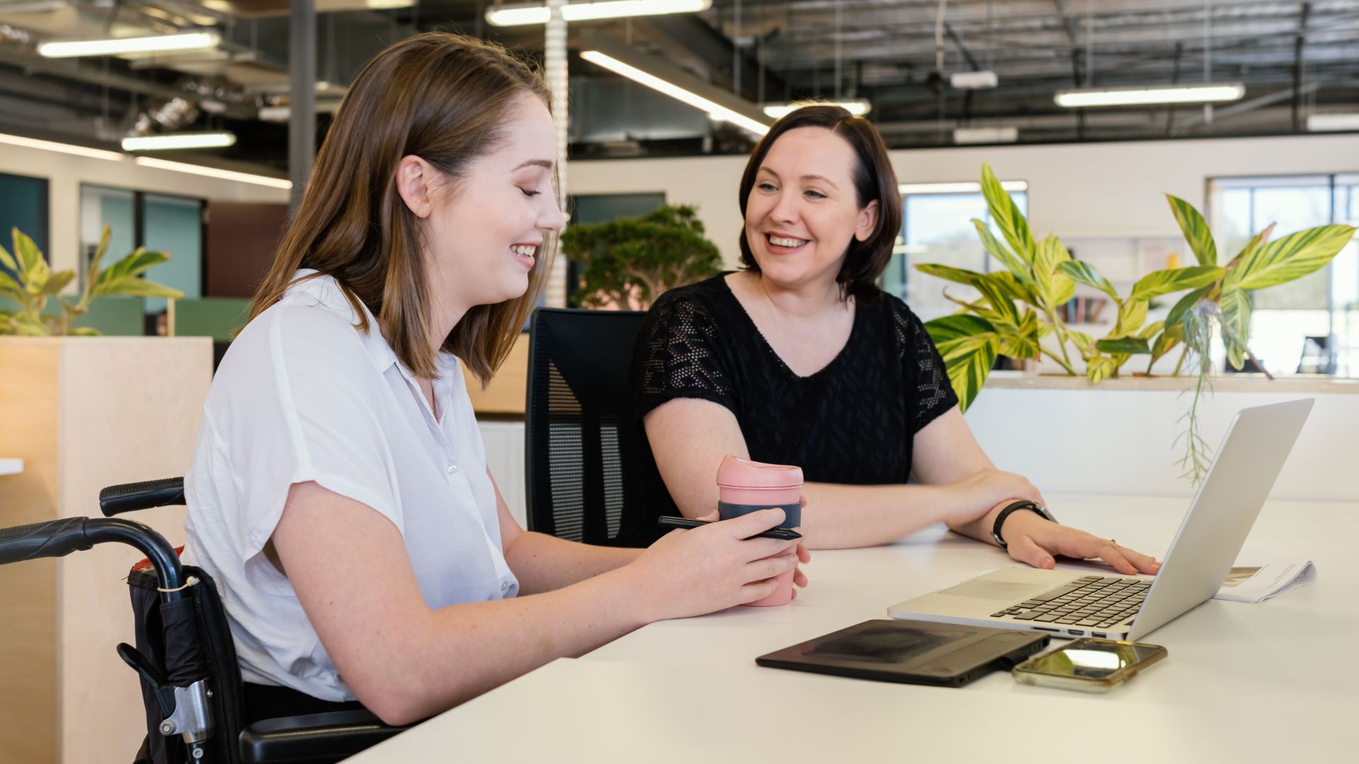 Two women sit together at a desk in a modern office, smiling and looking at a laptop. One woman uses a wheelchair and holds a reusable coffee cup, while the other offers support during a work discussion.