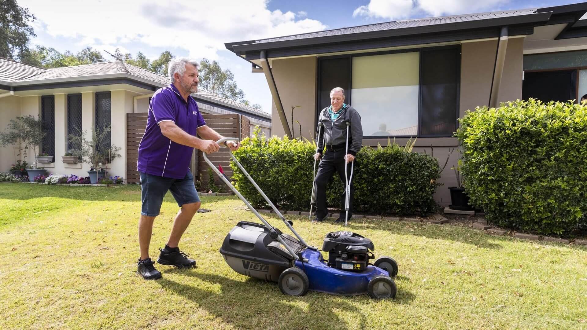 A man on crutches watching a man mow his front lawn