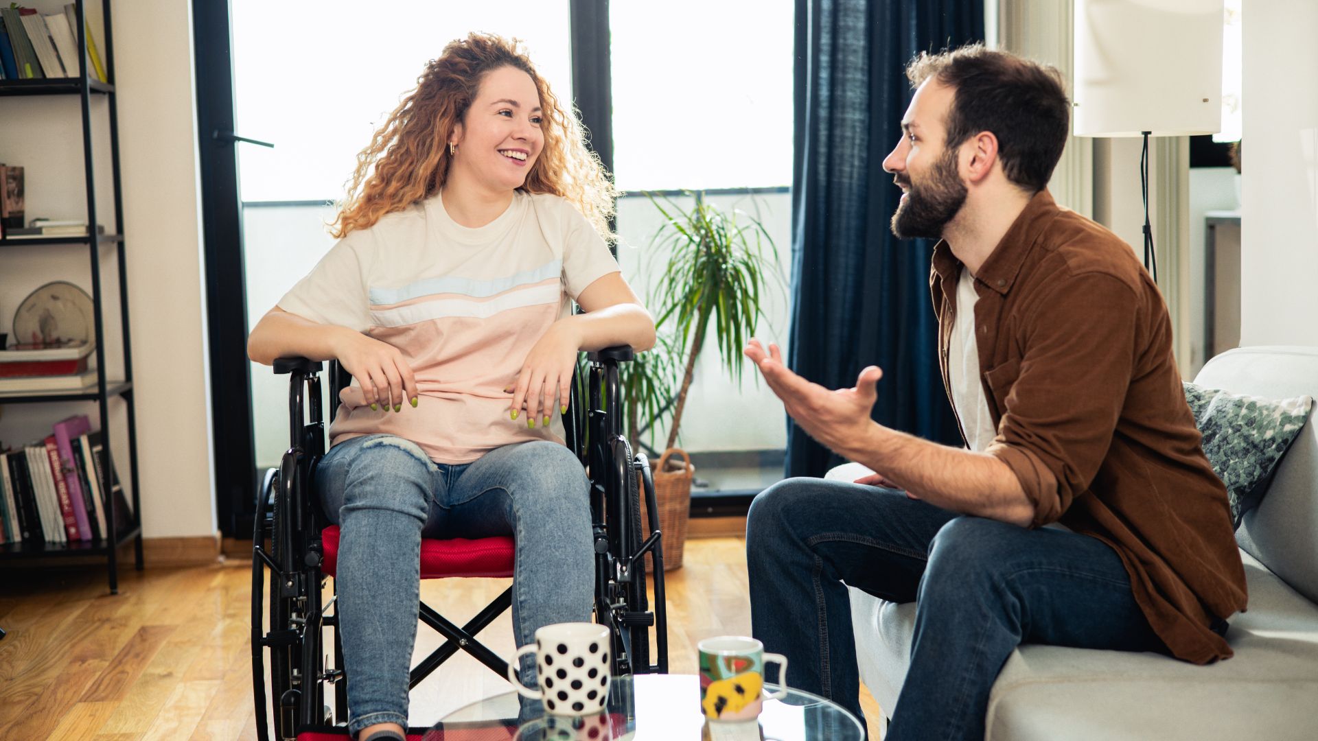 A woman using a wheelchair and a man sit facing each other in a living room, smiling and chatting comfortably, with natural light and indoor plants in the background.