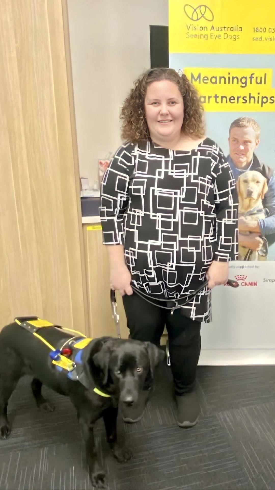Ayesha from APM Communities stands outside a public venue holding the lead of guide dog Princess, showing how guide dogs support safe navigation in community environments.