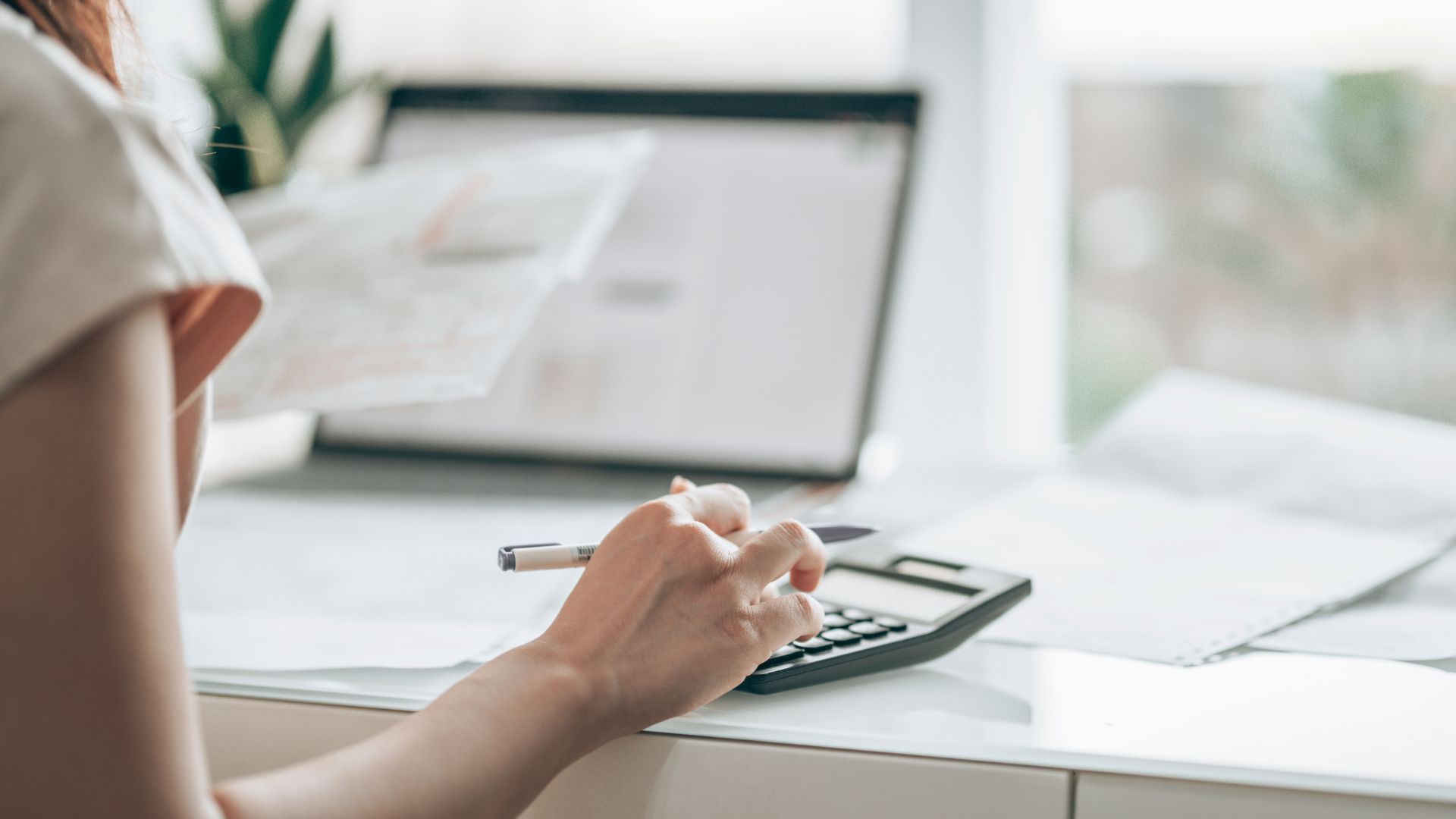 A person sits at a desk using a calculator and pen to review paperwork, with a laptop open in the background in a bright, modern workspace.