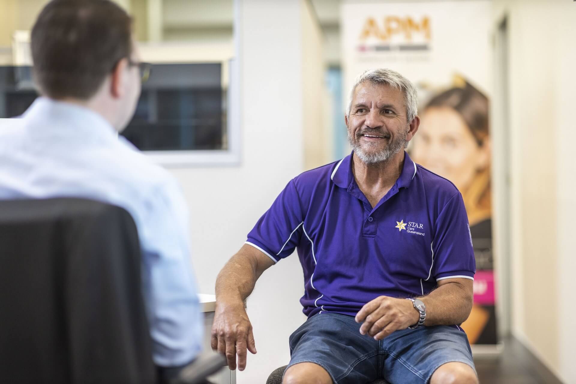 Jeff smiling in his purple uniform top leaning on an office wall