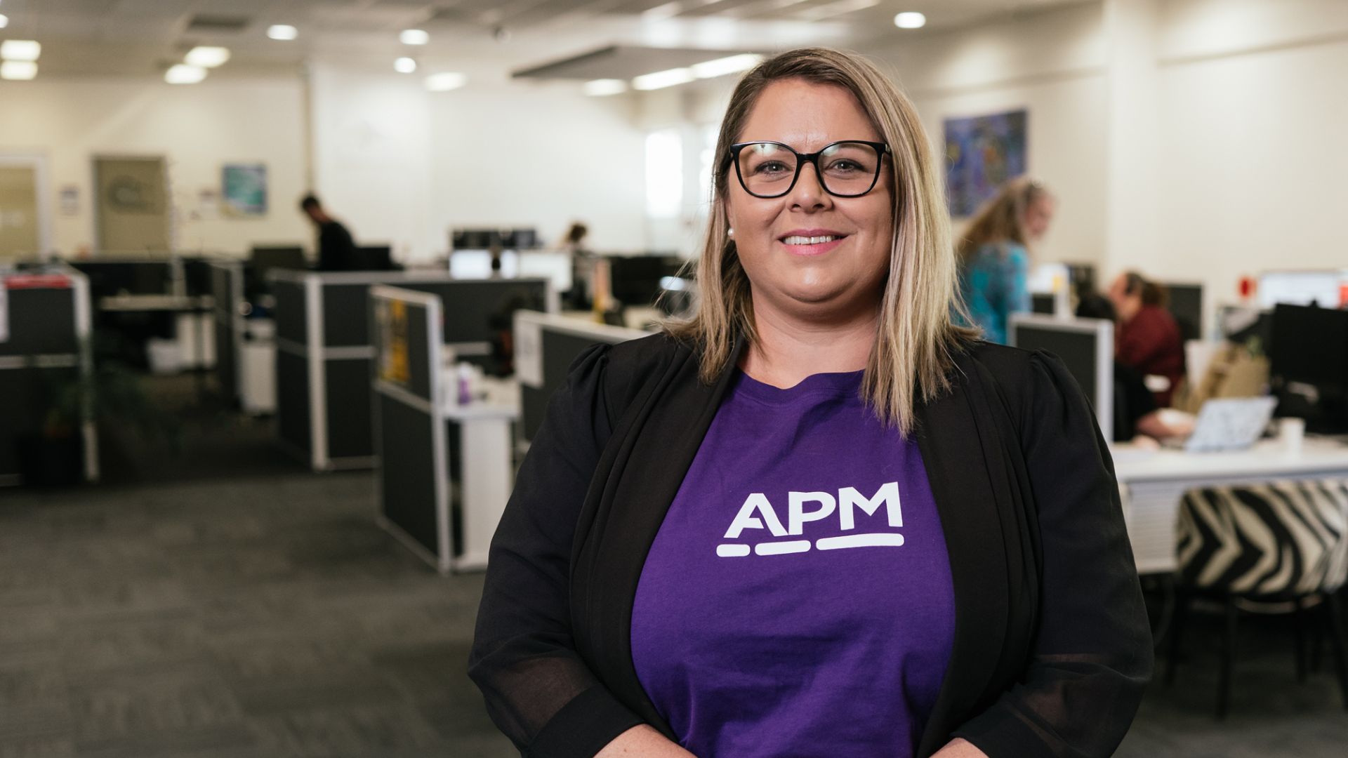 A woman in a purple APM top smiles in a local office