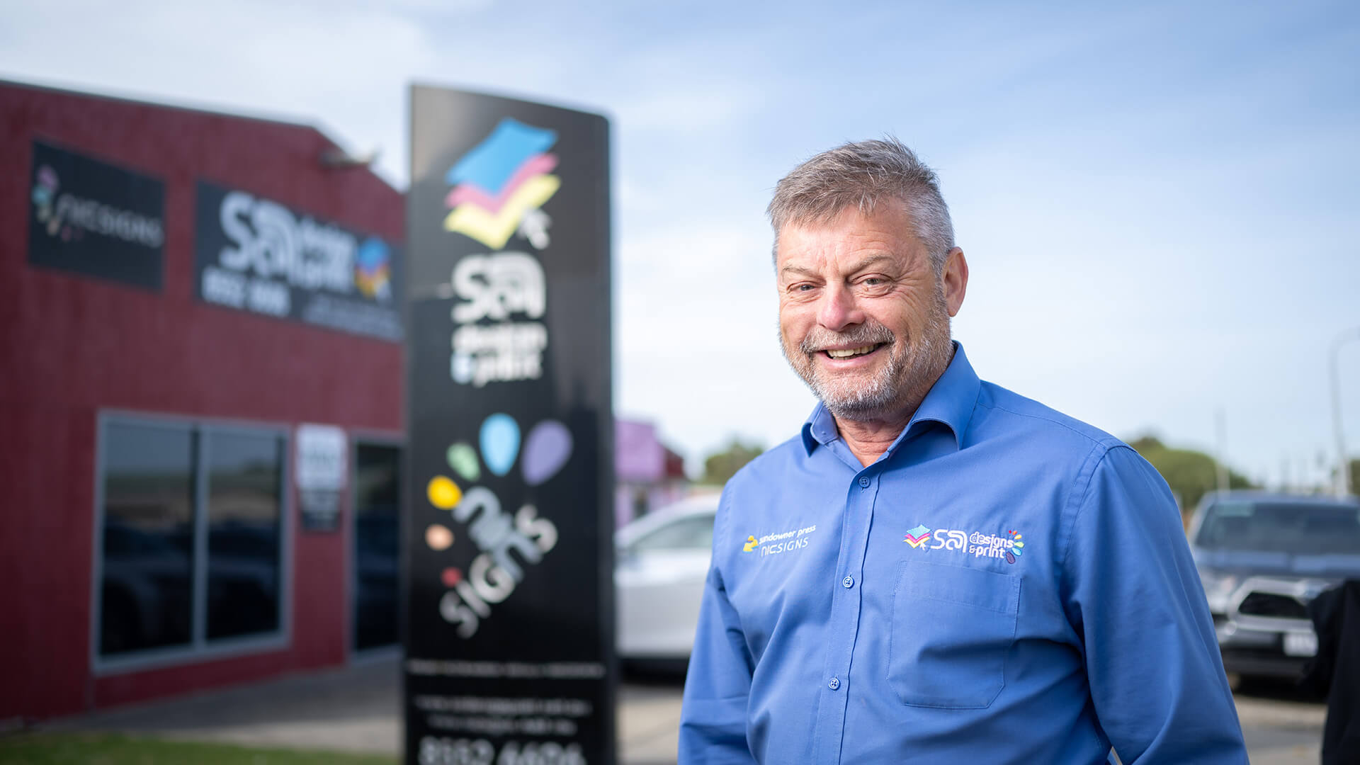 Man smiling in a blue shirt outside his business