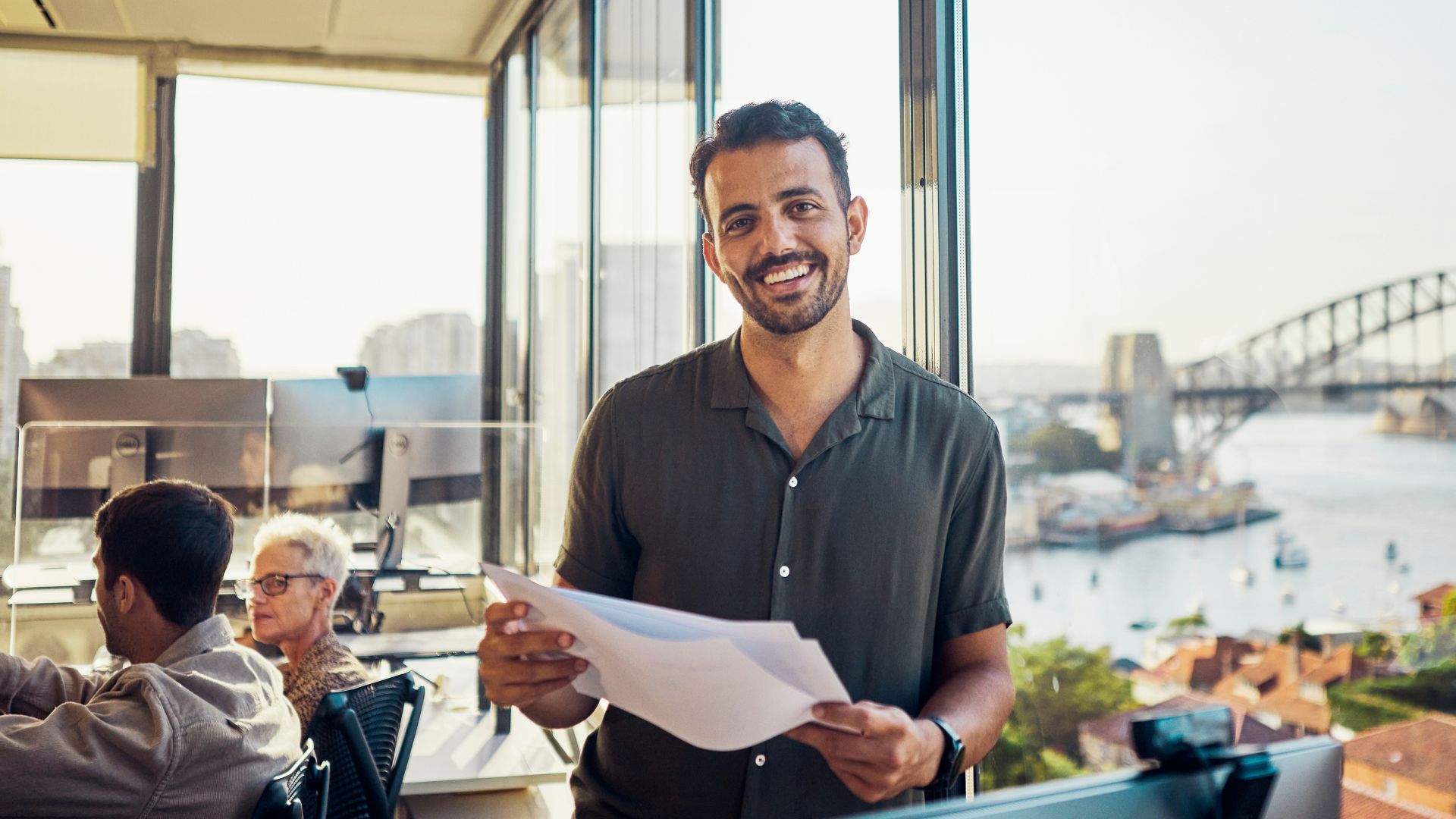 Man smiles holding paper in office overlooking Sydney Harbour