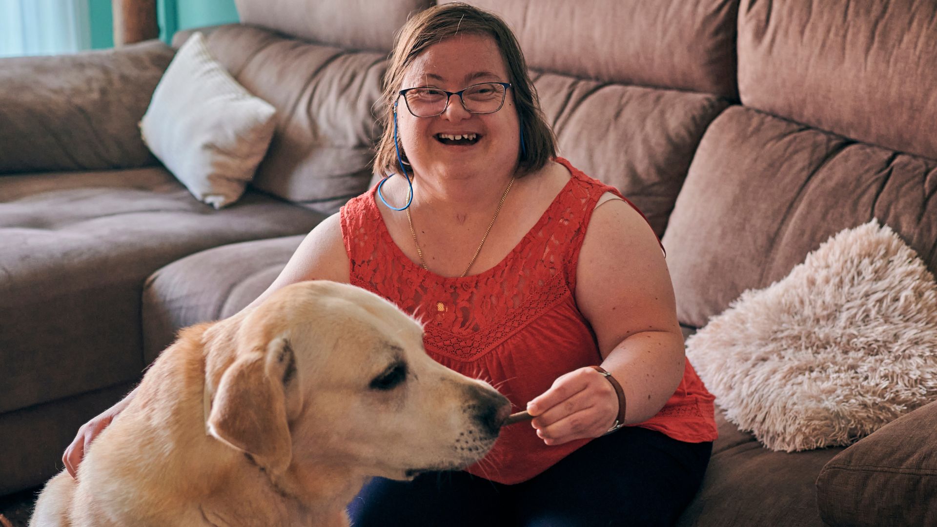A woman sits on a couch at home, smiling while offering a treat to a dog beside her, with soft furnishings and cushions in the background.