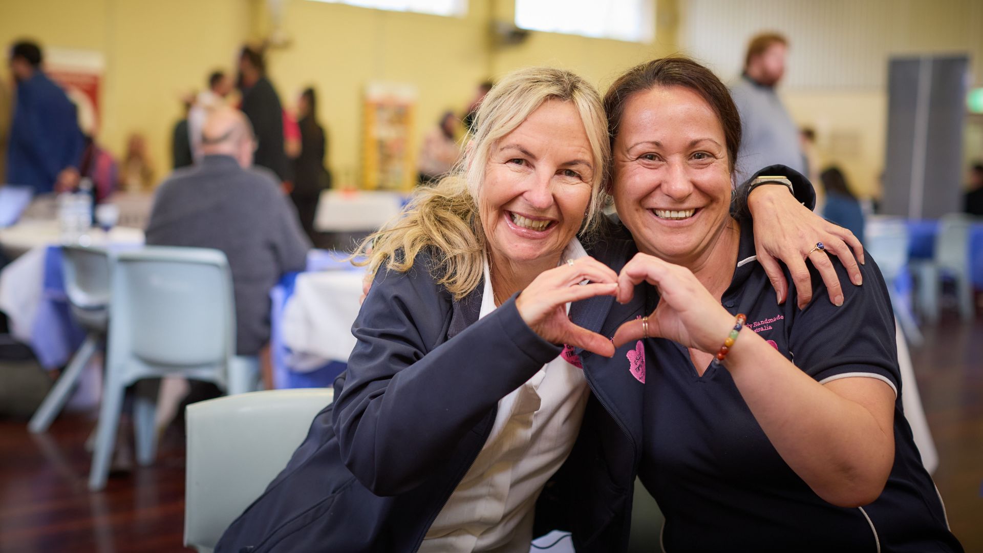 Two women make hearts with their hands in a community expo